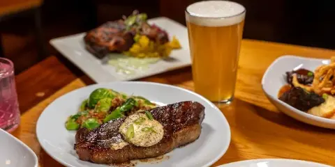 A table setting featuring a plate of steak with a butter topping, a glass of beer, and other prepared dishes.