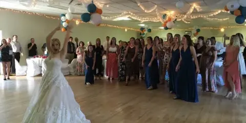A bride dances on a wooden floor surrounded by wedding guests in a decorated reception hall.