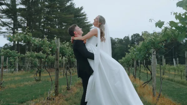 A groom in a black suit lifts a bride in a white gown in a vineyard.