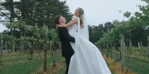 A groom in a black suit lifts a bride in a white gown in a vineyard.