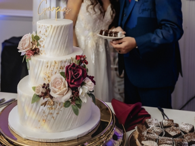 A multi-tiered white wedding cake with floral decorations on a gold stand, with a bride and groom standing behind it.