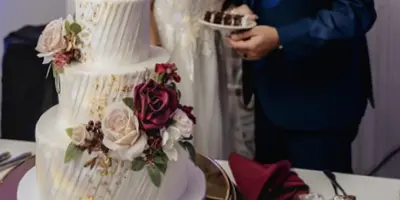 A multi-tiered white wedding cake with floral decorations on a gold stand, with a bride and groom standing behind it.