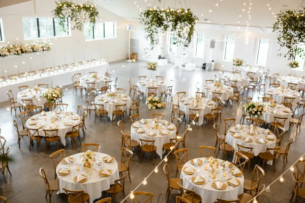 A spacious indoor wedding reception hall featuring round tables with white linens, floral centerpieces, and hanging string lights.