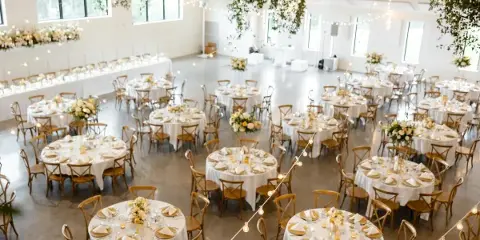 A spacious indoor wedding reception hall featuring round tables with white linens, floral centerpieces, and hanging string lights.