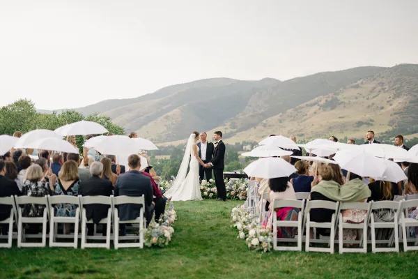 Outdoor wedding ceremony with bride and groom at the altar, guests seated in white chairs with umbrellas, floral aisle, and mountain backdrop.
