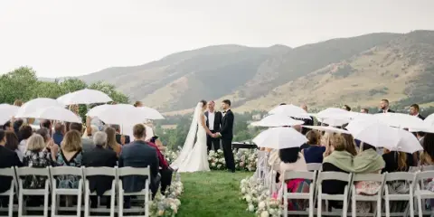 Outdoor wedding ceremony with bride and groom at the altar, guests seated in white chairs with umbrellas, floral aisle, and mountain backdrop.