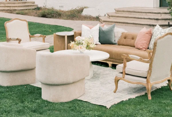 An outdoor lounge area featuring a tan leather sofa, cream armchairs, a round white coffee table with flowers, and a patterned rug on a lawn.