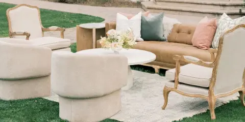An outdoor lounge area featuring a tan leather sofa, cream armchairs, a round white coffee table with flowers, and a patterned rug on a lawn.