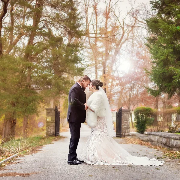 A bride and groom standing face-to-face on a paved path surrounded by trees and autumn foliage.