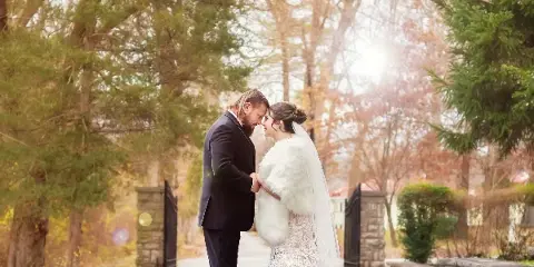 A bride and groom standing face-to-face on a paved path surrounded by trees and autumn foliage.