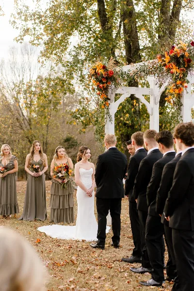 A bride and groom standing at a floral-decorated arch during an outdoor wedding ceremony with their wedding party.