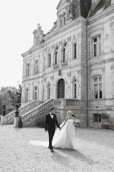 Black and white photo of a bride and groom walking hand in hand in front of a large stone building.