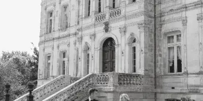 Black and white photo of a bride and groom walking hand in hand in front of a large stone building.
