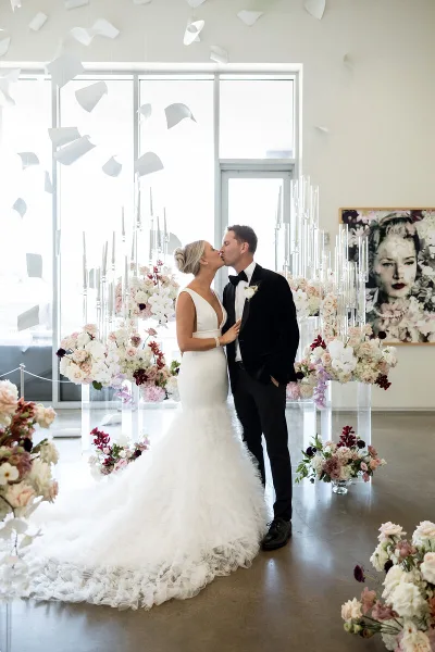 A bride and groom kissing in a modern venue surrounded by elaborate floral arrangements and tall candle displays.