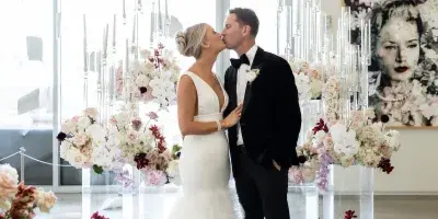 A bride and groom kissing in a modern venue surrounded by elaborate floral arrangements and tall candle displays.