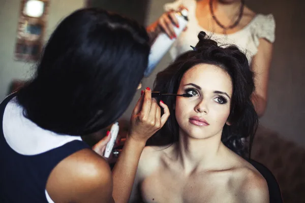 A makeup artist applying mascara to a bride while a hairstylist works on her hair in the background.