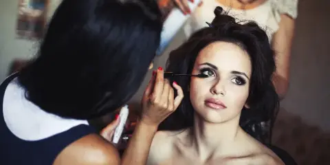 A makeup artist applying mascara to a bride while a hairstylist works on her hair in the background.