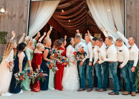 A wedding party posing and celebrating in front of a rustic wooden barn entrance decorated with white drapery and string lights.