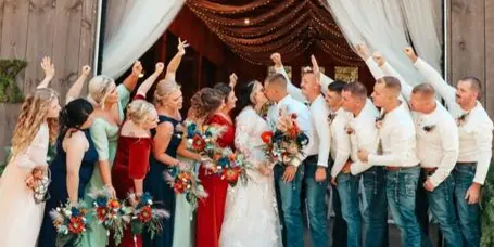 A wedding party posing and celebrating in front of a rustic wooden barn entrance decorated with white drapery and string lights.