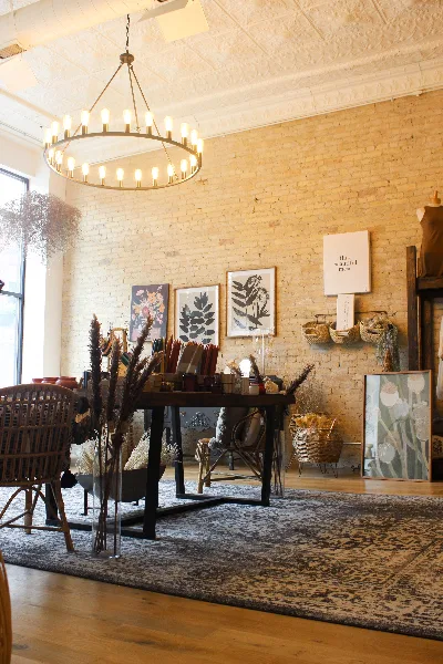 Interior view of a boutique shop with a large chandelier, brick walls, floral art prints, and a display table with dried flowers and home goods.