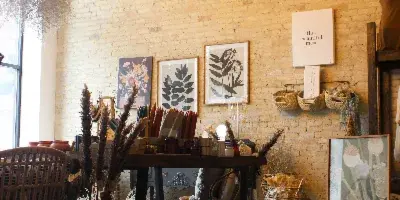Interior view of a boutique shop with a large chandelier, brick walls, floral art prints, and a display table with dried flowers and home goods.