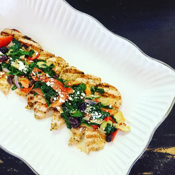 Plated grilled chicken topped with spinach, tomatoes, olives, and crumbled cheese on a white serving dish.