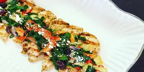 Plated grilled chicken topped with spinach, tomatoes, olives, and crumbled cheese on a white serving dish.