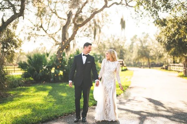 A bride and groom walking hand-in-hand on a path outdoors, surrounded by trees and greenery.