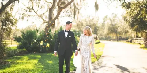 A bride and groom walking hand-in-hand on a path outdoors, surrounded by trees and greenery.