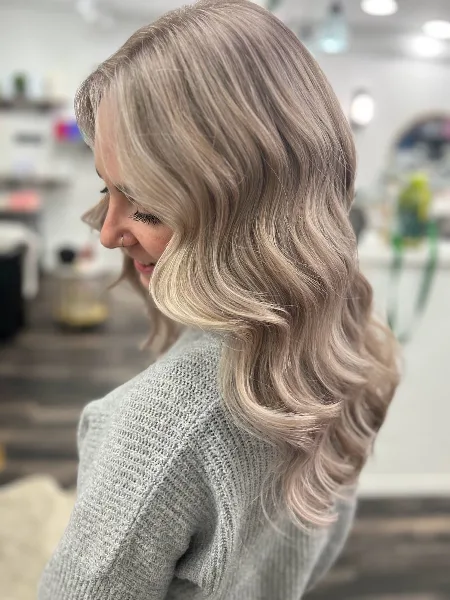 A woman with styled, wavy blonde hair shown in profile within a salon environment.