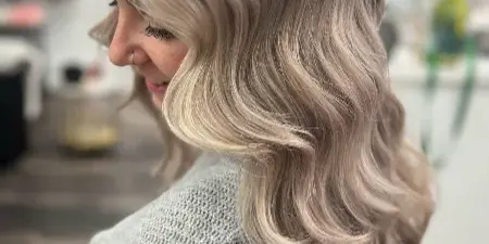 A woman with styled, wavy blonde hair shown in profile within a salon environment.
