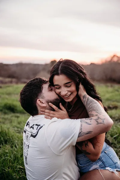A professional outdoor engagement photograph of a smiling couple embracing in a grassy field at sunset.