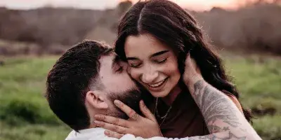 A professional outdoor engagement photograph of a smiling couple embracing in a grassy field at sunset.