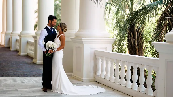 A bride and groom standing together in a white columned corridor, with the bride holding a bouquet of white flowers.