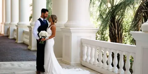 A bride and groom standing together in a white columned corridor, with the bride holding a bouquet of white flowers.