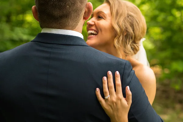 A bride in a white dress and veil smiles at a groom in a dark suit, her hand with a ring resting on his shoulder against a green outdoor background.