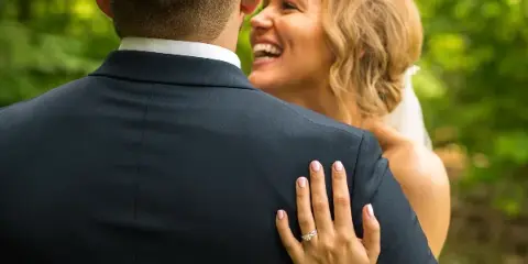 A bride in a white dress and veil smiles at a groom in a dark suit, her hand with a ring resting on his shoulder against a green outdoor background.