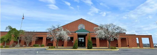 Exterior view of a red brick community hall with a central entrance, flagpole, and parking lot under a blue sky.