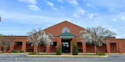 Exterior view of a red brick community hall with a central entrance, flagpole, and parking lot under a blue sky.