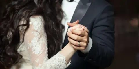 Close-up of a bride and groom holding hands, showing the bride's lace sleeve and the groom's suit.