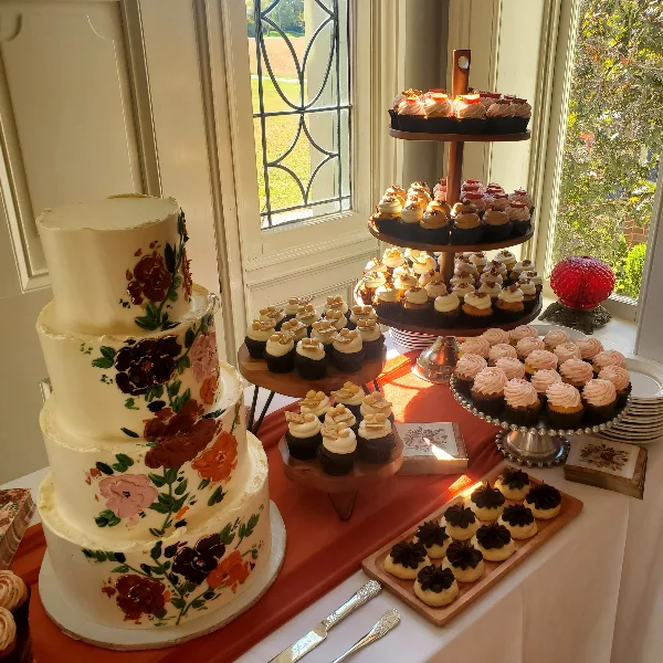 A four-tier white wedding cake with hand-painted floral designs sits next to a tiered stand of cupcakes and a tray of small pastries on a table near a window.