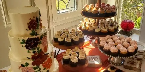 A four-tier white wedding cake with hand-painted floral designs sits next to a tiered stand of cupcakes and a tray of small pastries on a table near a window.