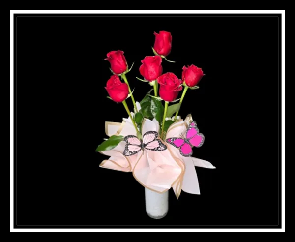 A bouquet of six red roses arranged in a white designer vase with decorative pink butterflies.