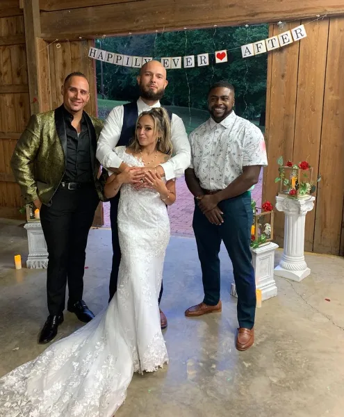 A wedding couple posing with two entertainment professionals at a rustic barn venue with a 'Happily Ever After' banner.