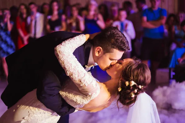 A bride and groom sharing a dip during a dance at a wedding reception, surrounded by guests and illuminated by colored event lighting.