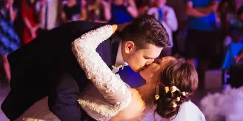 A bride and groom sharing a dip during a dance at a wedding reception, surrounded by guests and illuminated by colored event lighting.