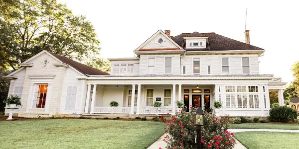 Exterior view of a historic white two-story building with a columned porch, manicured lawn, and flowering shrubs, representing a wedding and event venue.