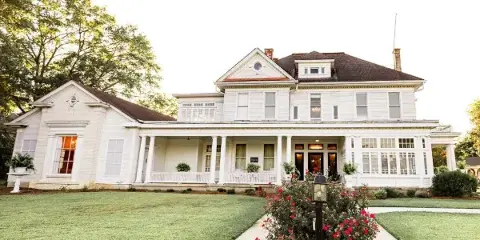 Exterior view of a historic white two-story building with a columned porch, manicured lawn, and flowering shrubs, representing a wedding and event venue.