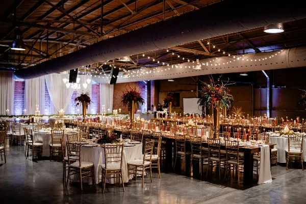 Interior view of an industrial-chic wedding venue featuring exposed brick walls, high ceilings with string lights, and round tables set with white linens and gold chiavari chairs.