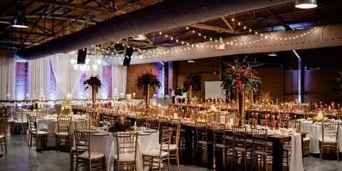 Interior view of an industrial-chic wedding venue featuring exposed brick walls, high ceilings with string lights, and round tables set with white linens and gold chiavari chairs.